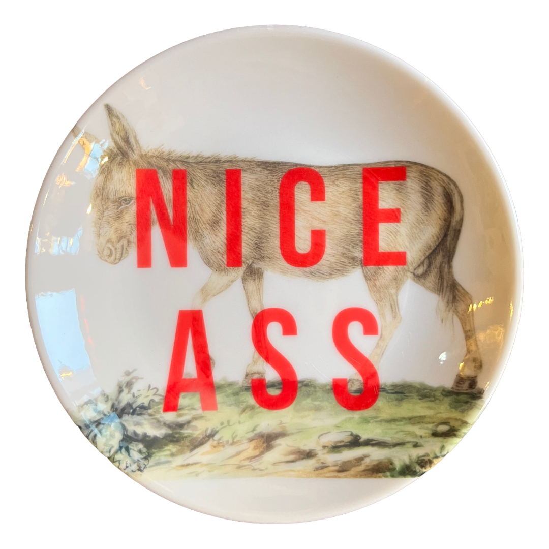 A round white ceramic plate featuring a printed illustration of a donkey in profile, with the words 'NICE ASS' printed in large red letters across the center.