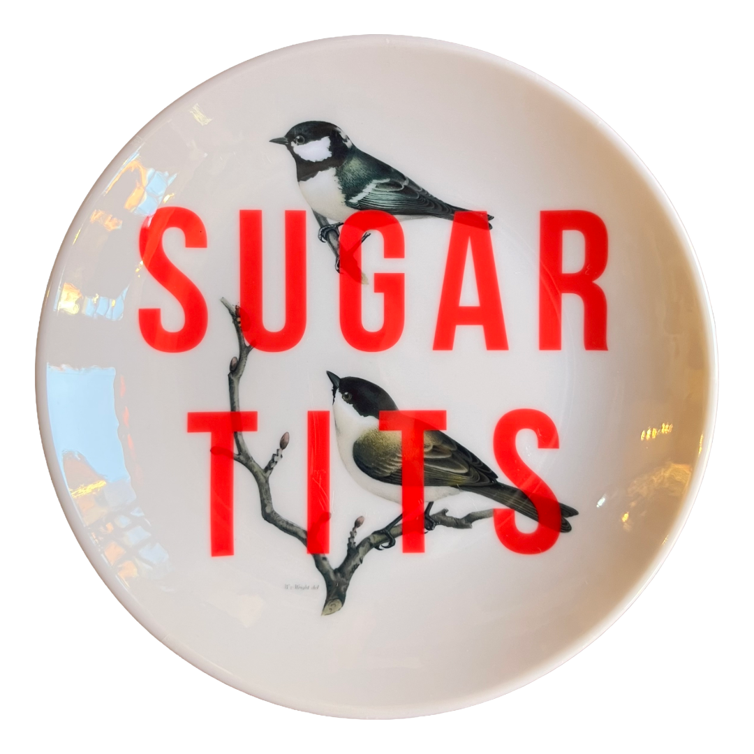 A round white ceramic plate featuring a printed design of two birds perched on a branch, with the words 'SUGAR TITS' in large red letters across the front.