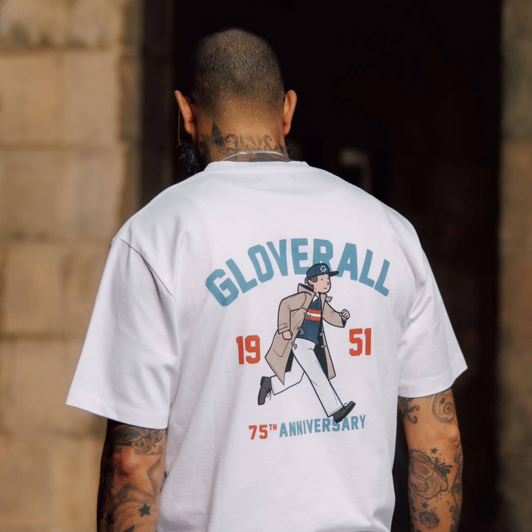 A man viewed from behind wearing a white Gloover 75th Anniversary t-shirt with a large graphic print on the back, set against a dark doorway and stone wall background.