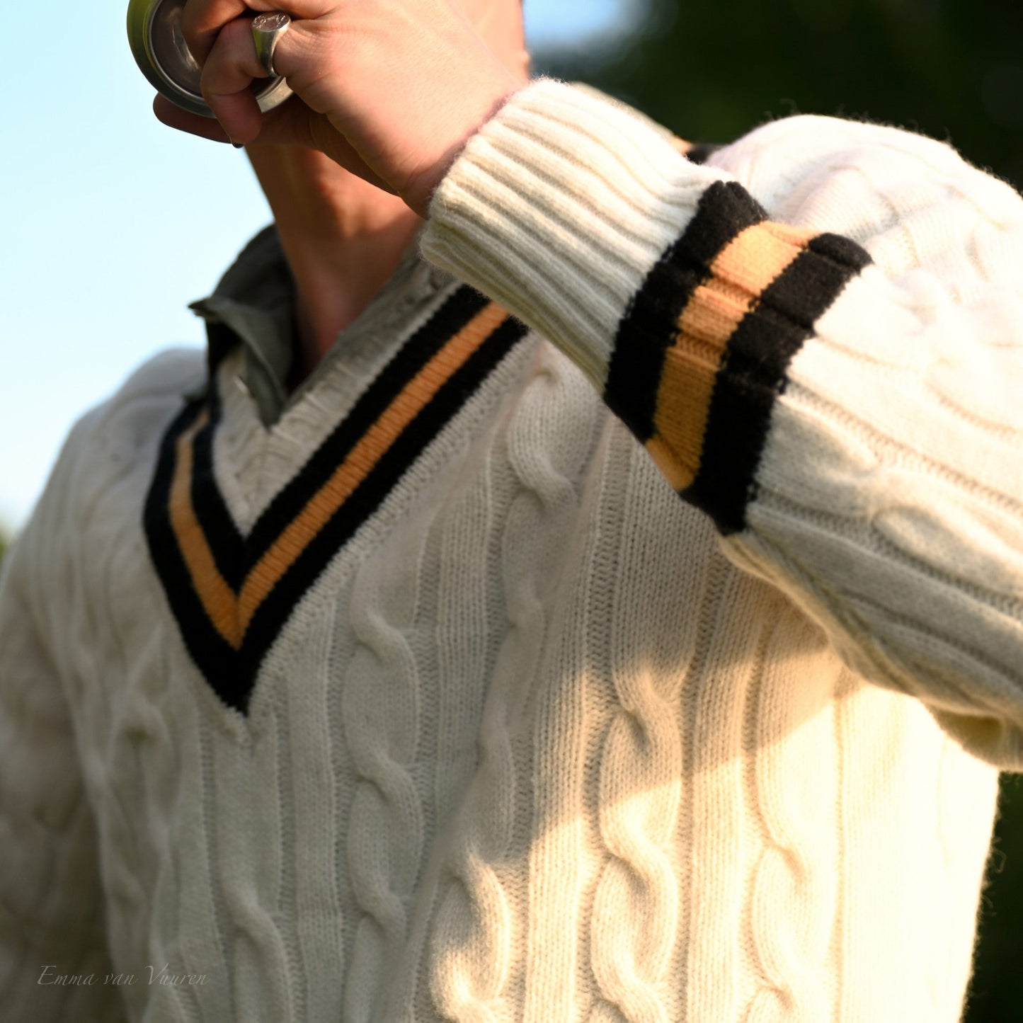 Person wearing a white sweater with black and yellow stripes, drinking from a can outdoors.