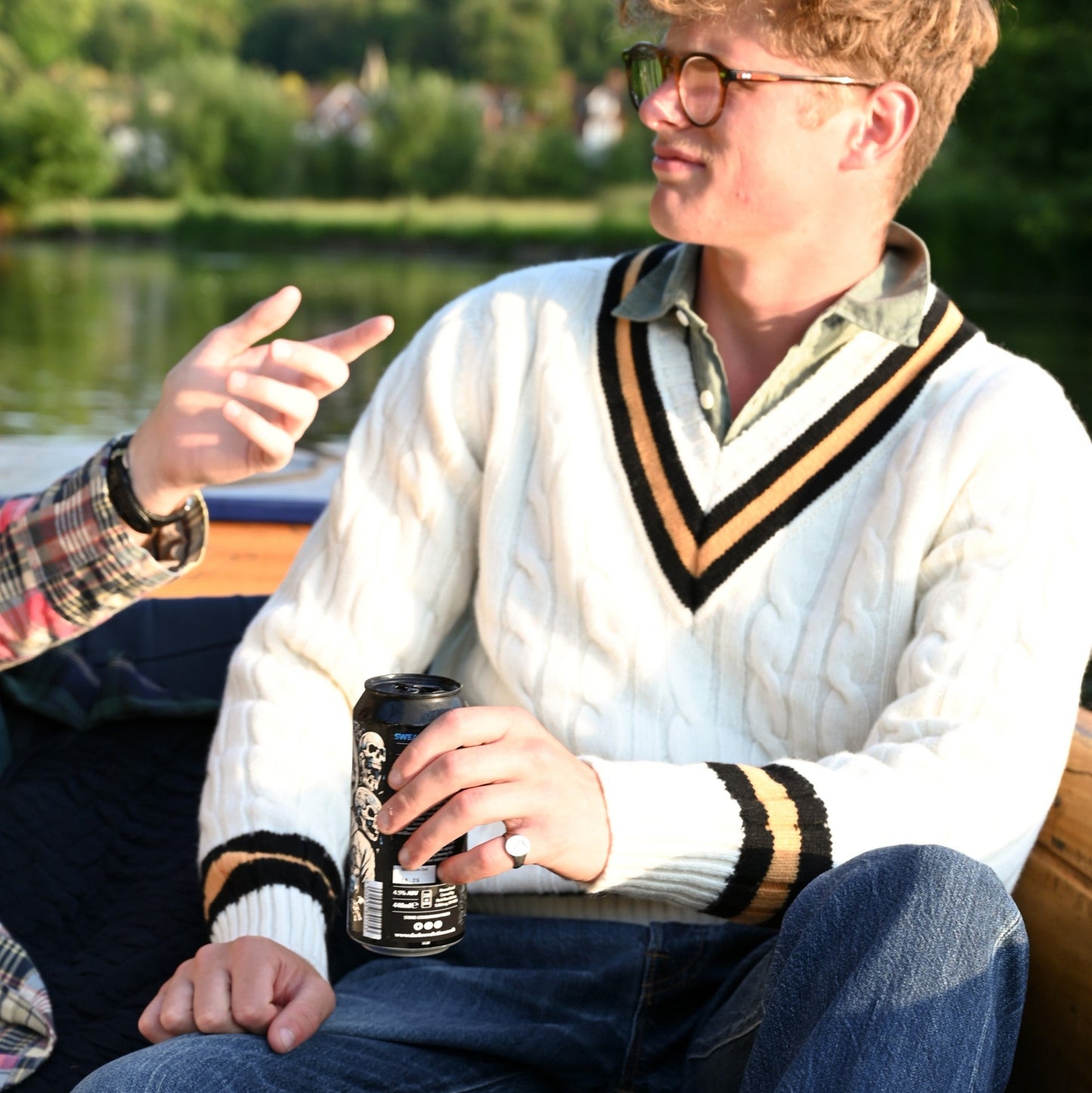 Person sitting by a lake, wearing a white sweater with black and gold trim, holding a can.