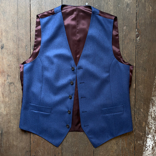 A denim blue wool flannel waistcoat with a V-neck collar, buttoned front, and welted pockets, displayed on a wooden surface.