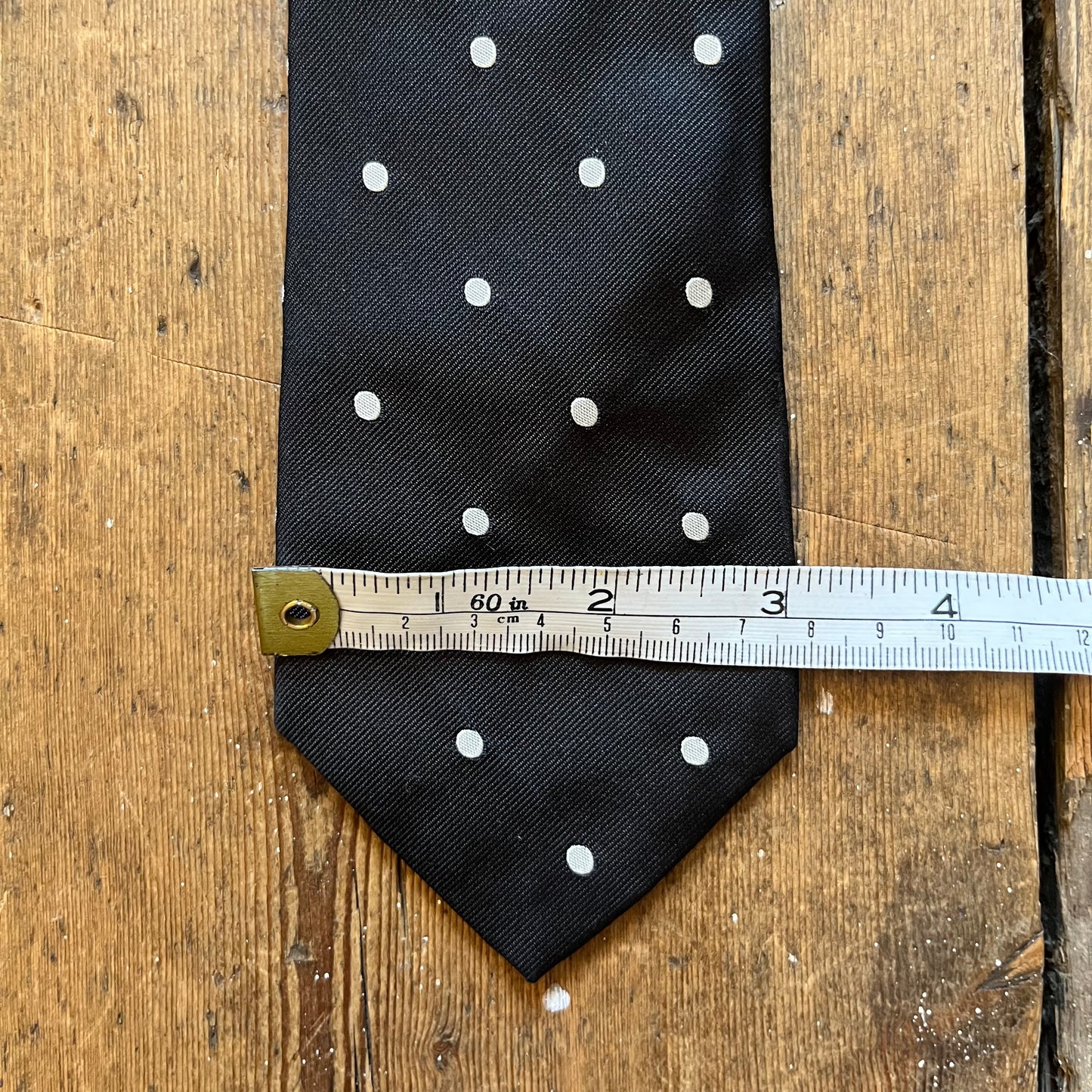 A black necktie with white polka dots laid flat on a rustic wooden surface, with a metal tape measure placed horizontally across the middle to show the size.