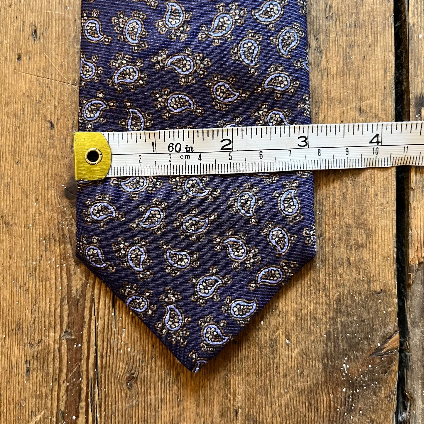 A navy blue necktie with a white and gold paisley pattern, laid flat on a rustic wooden surface and measured with a metal tape measure.