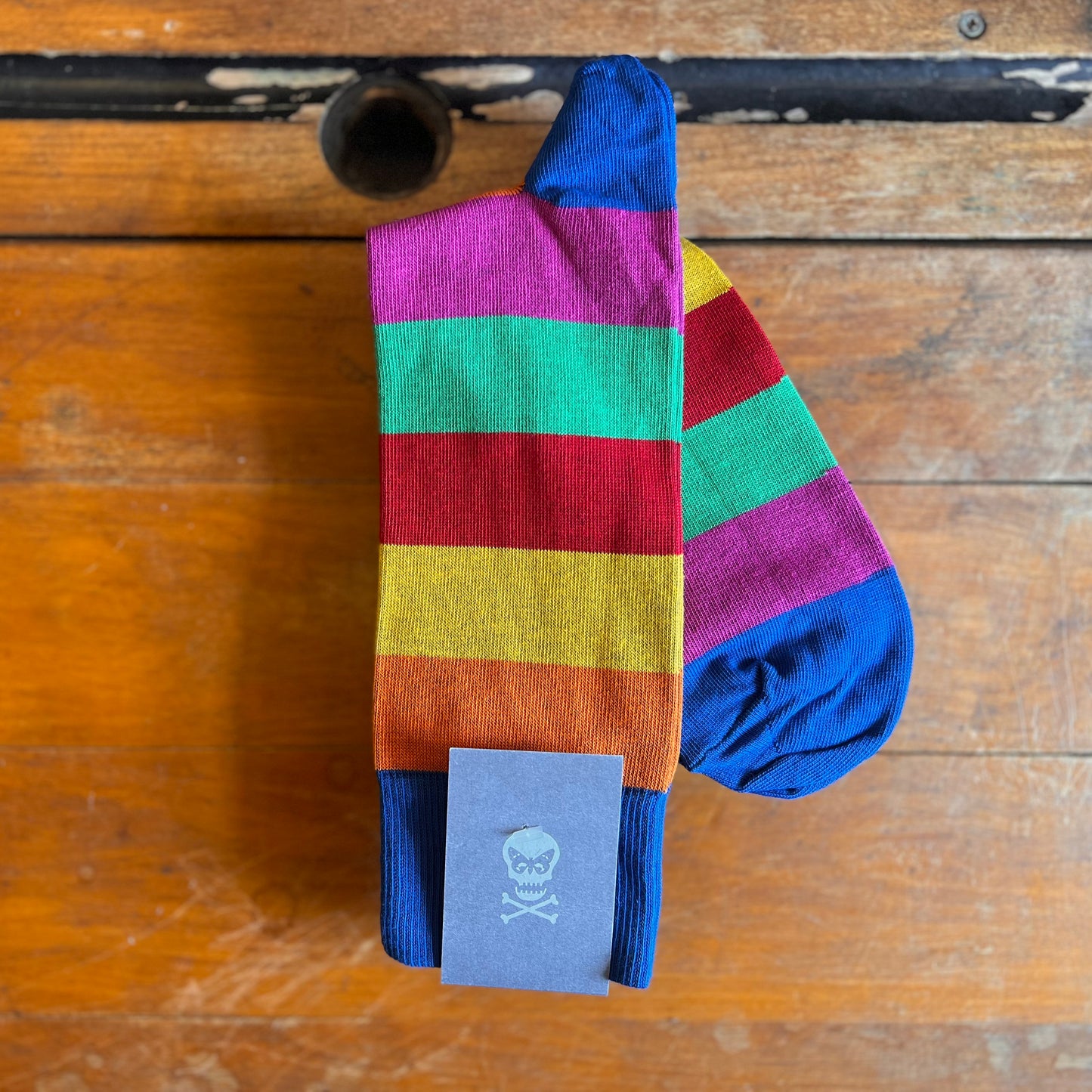 A pair of socks with stripes sit atop a wooden surface. The stripes are Orange, yellow, red, green and pink. The top, heel and toe of the sock are blue.