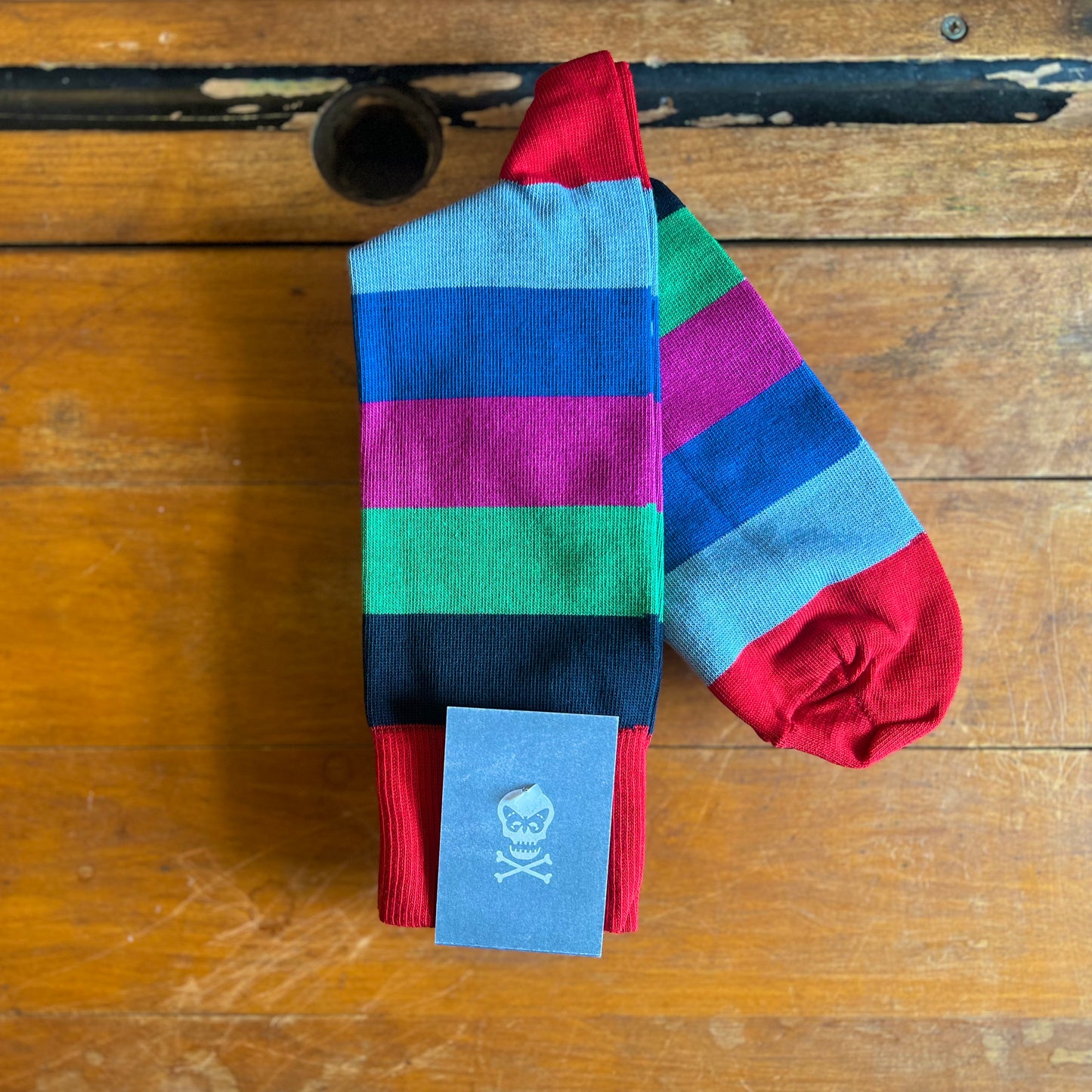 A multi coloured striped sock sits on a wooden backdrop. The stripes are red, navy, green, pink, blue and azure.