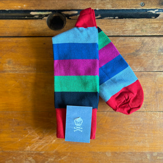 A multi coloured striped sock sits on a wooden backdrop. The stripes are red, navy, green, pink, blue and azure.