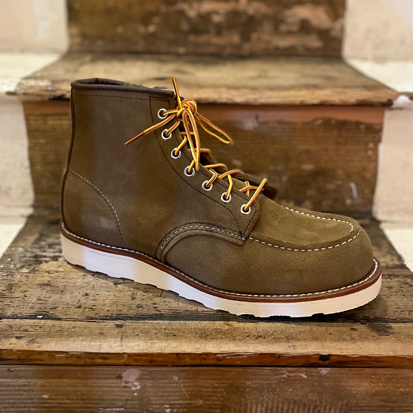 A brown olive mohave suede Red Wing classic moc-toe boot with white traction tread sole and leather footbed, displayed on a wooden step.