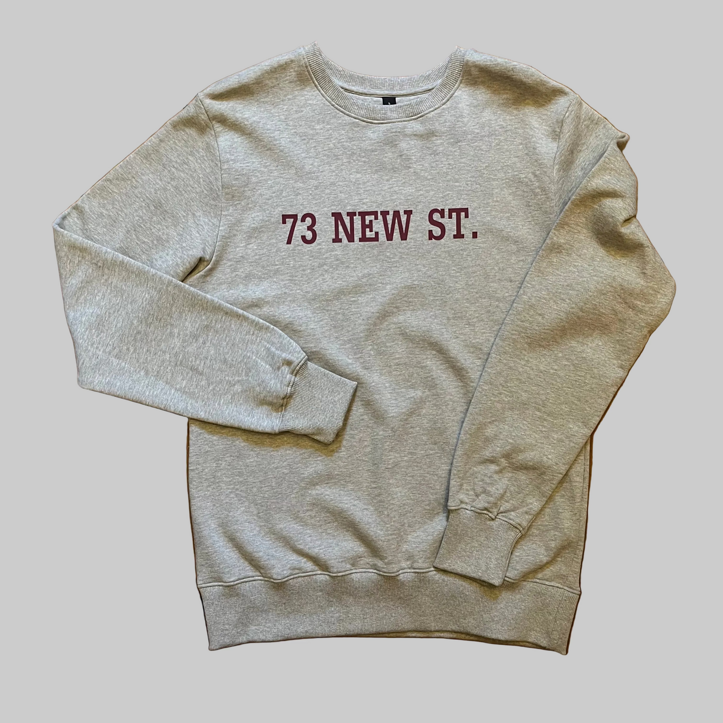 Grey cotton sweatshirt with '73 New Street' in red collegiate writing on the chest, placed on wooden boards
