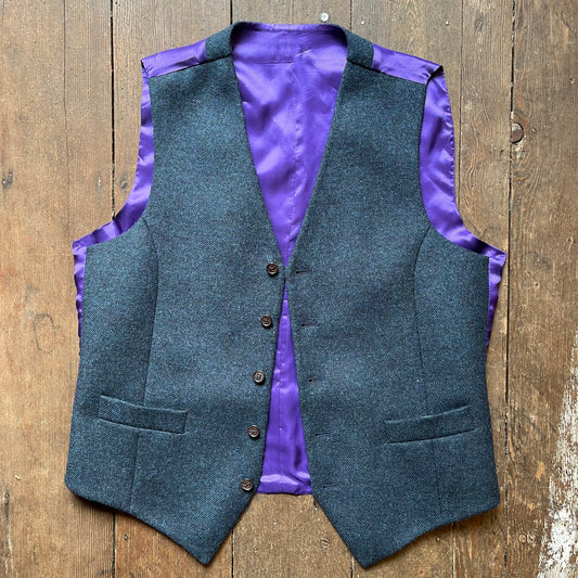 A blue tweed waistcoat with a peacock blue tone, featuring a five-button front, welted pockets, and a purple silk-like lining. It is laid out flat on a wooden surface.