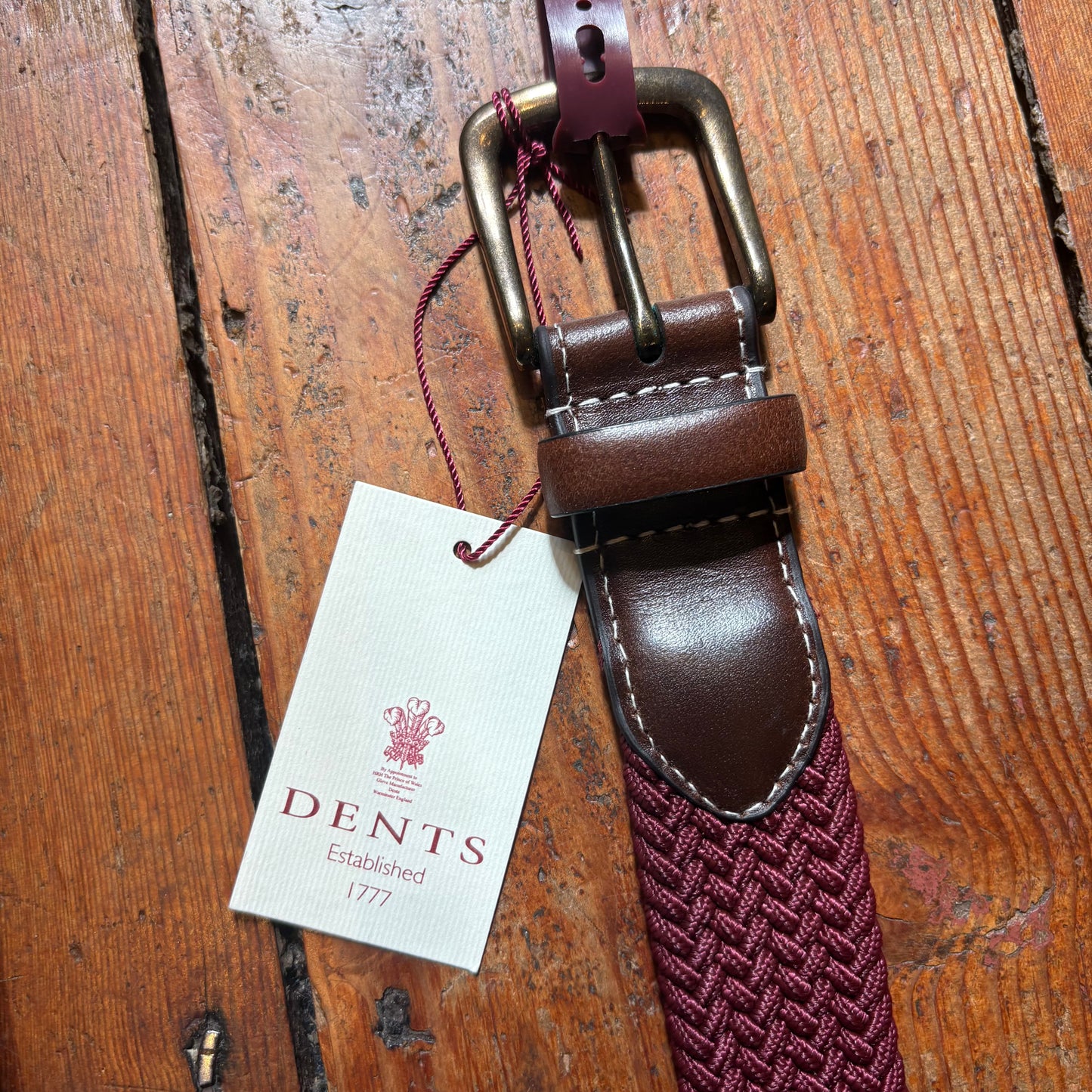 Brown leather belt with a maroon woven strap and Dents label on a wooden surface