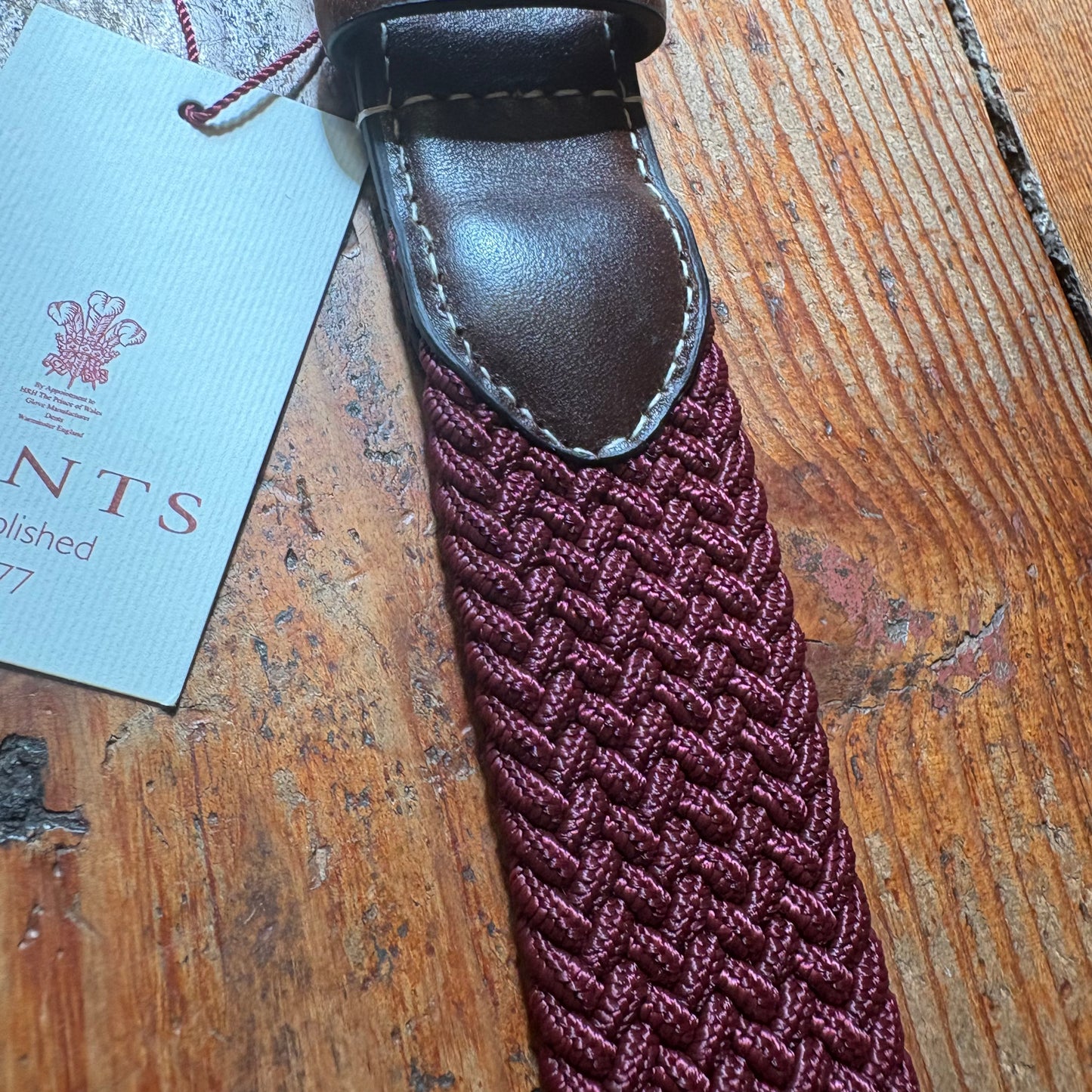 Maroon braided belt with leather buckle on a wooden surface, accompanied by a blue tag.