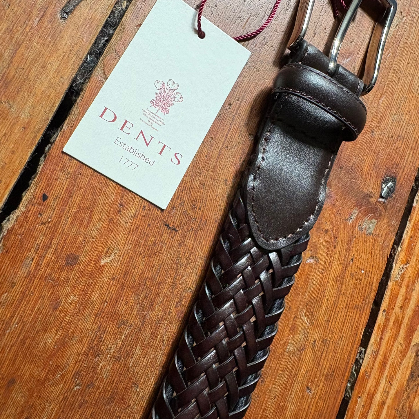 Brown leather belt with woven pattern on a wooden surface, featuring a Dents branded tag.