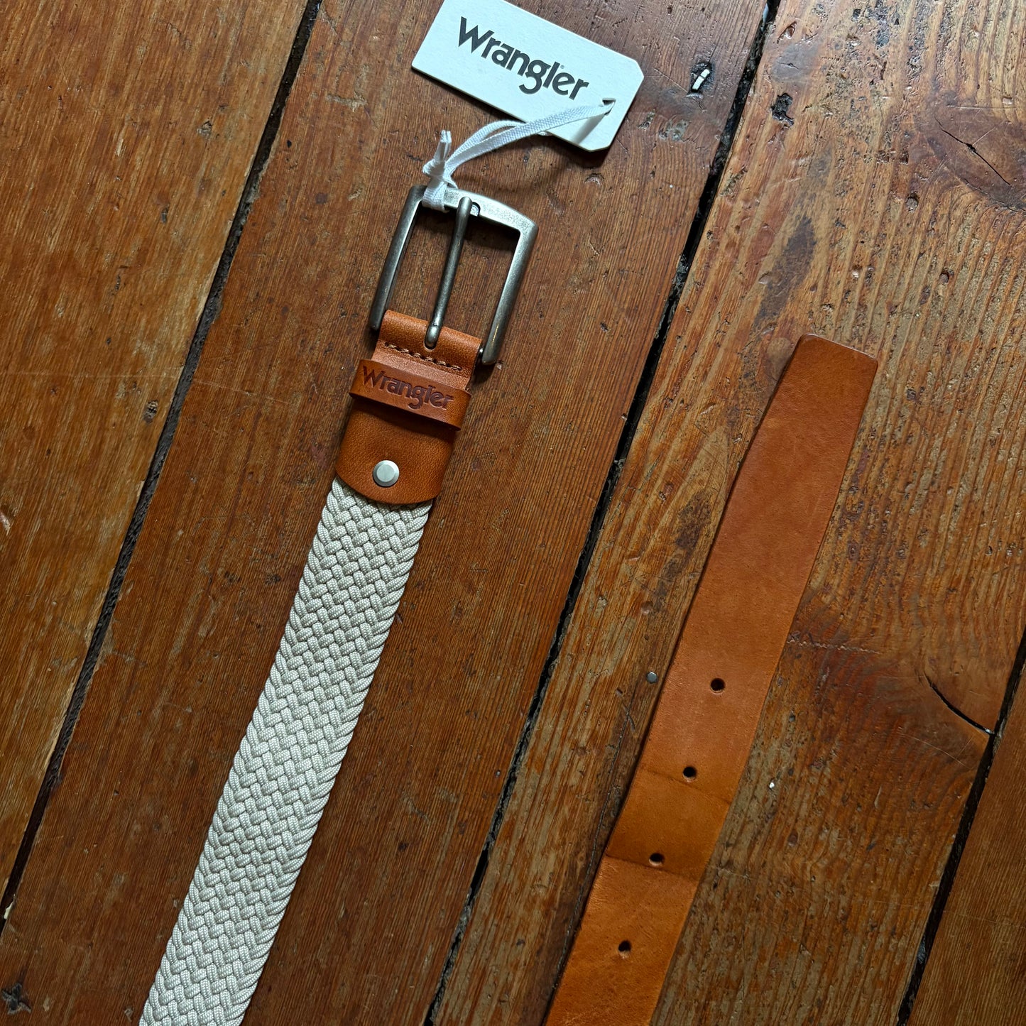 Two belts, one beige and one brown, with 'Wrangler' branding on a wooden surface.