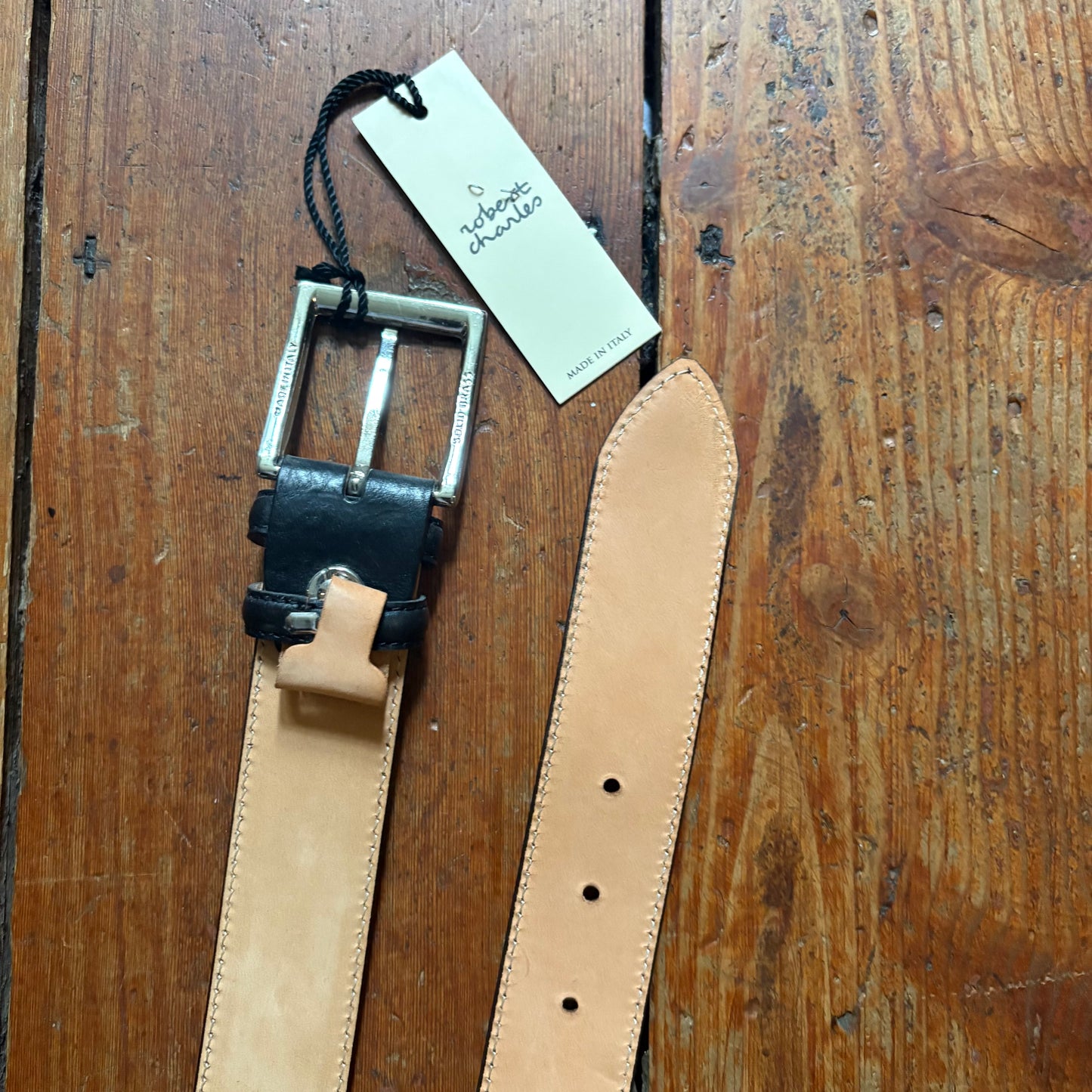 Leather belt with black buckle on a wooden surface, featuring a tag.