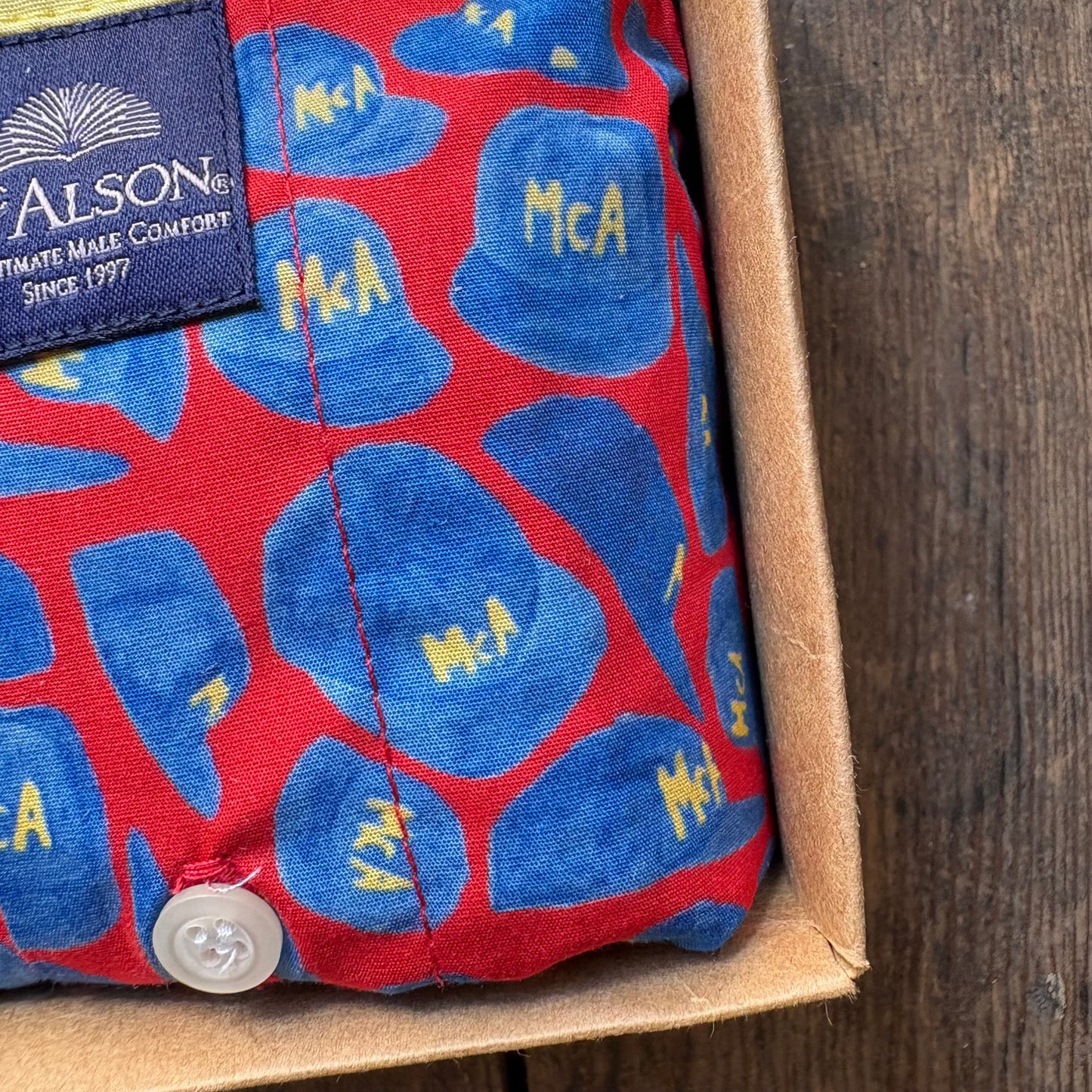 A close-up of a red and blue patterned fabric pouch with yellow 'MCA' lettering, a visible Nelson Alson brand label, and a white button, sitting inside a brown cardboard box