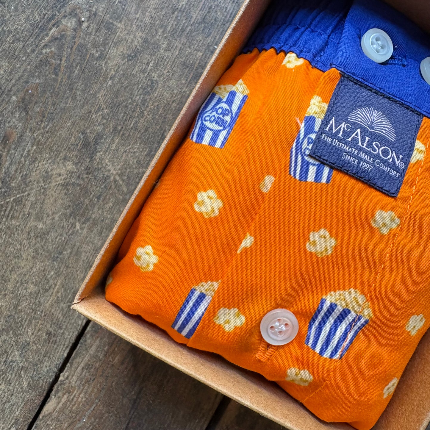 An orange children's button-up shirt by McAlson The Distinctive Kids Company, featuring a popcorn and blue striped pattern, folded inside a cardboard box on a rustic wooden surface.