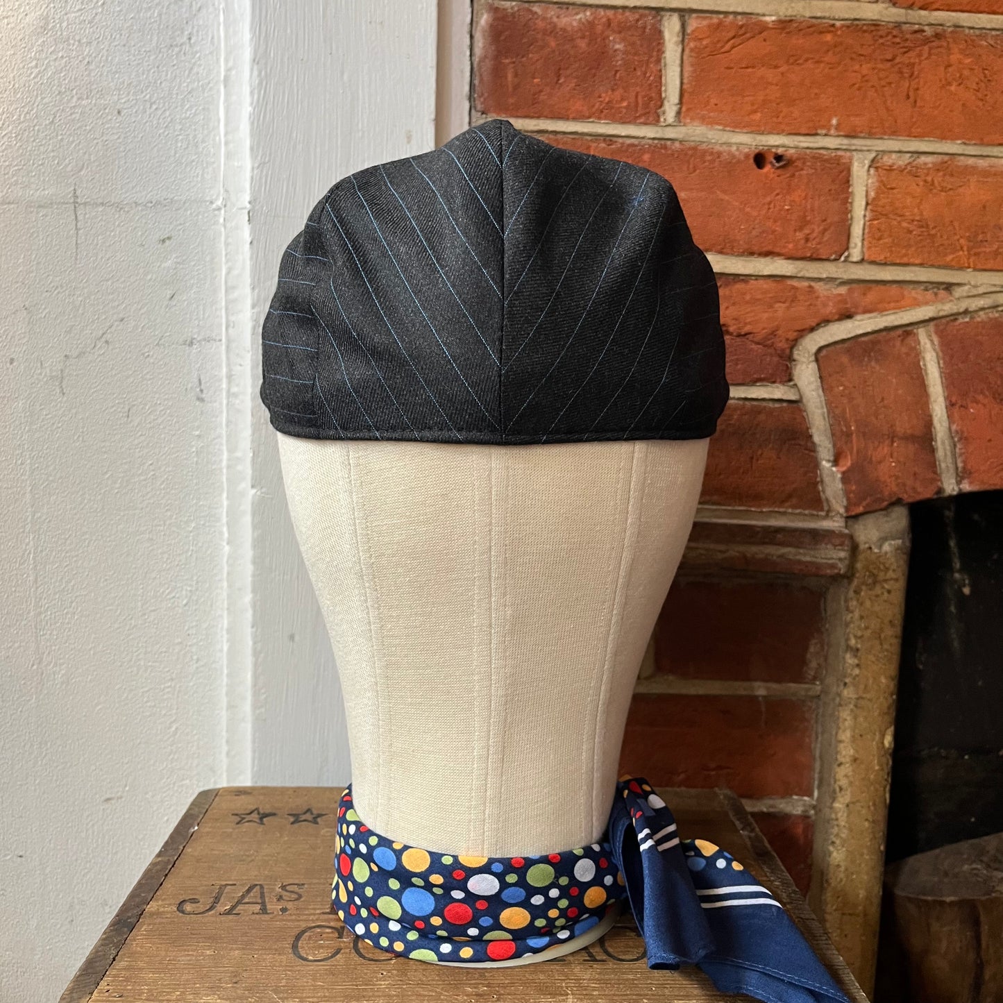Black pinstripe cap on a mannequin head with a polka dot band against a brick wall.