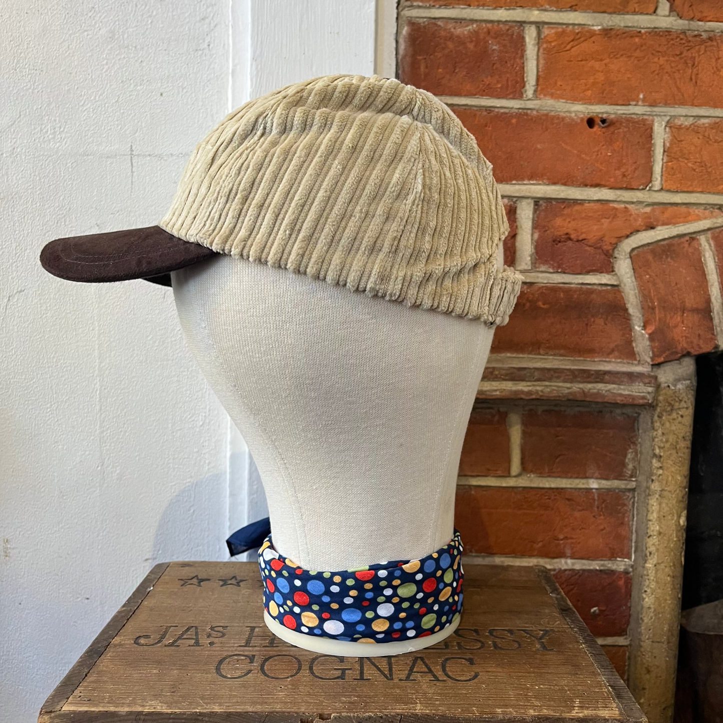 Mannequin head wearing a beige cap with a colorful bandana around its neck, against a brick wall.