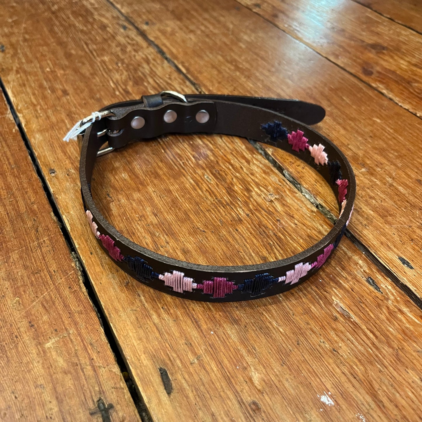 A brown leather dog collar with silver-style buckle fastening and a D-ring for lead attachment, featuring a woven pampa cross design in pink and navy colors.