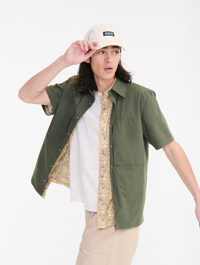 Person wearing a green shirt over a white and gold patterned shirt, with a beige cap.