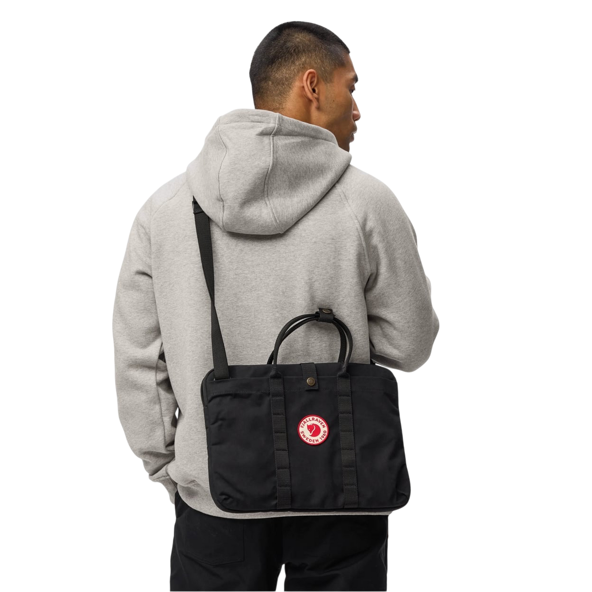 A man wearing a light gray hoodie and black pants, shown from the back while wearing a black laptop bag with a red circular logo on the back.