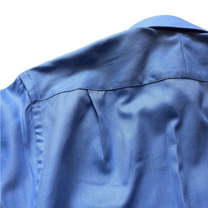Close-up of the back of a blue textured button-up shirt, showing the collar seam, shoulder stitching, and fine woven fabric texture