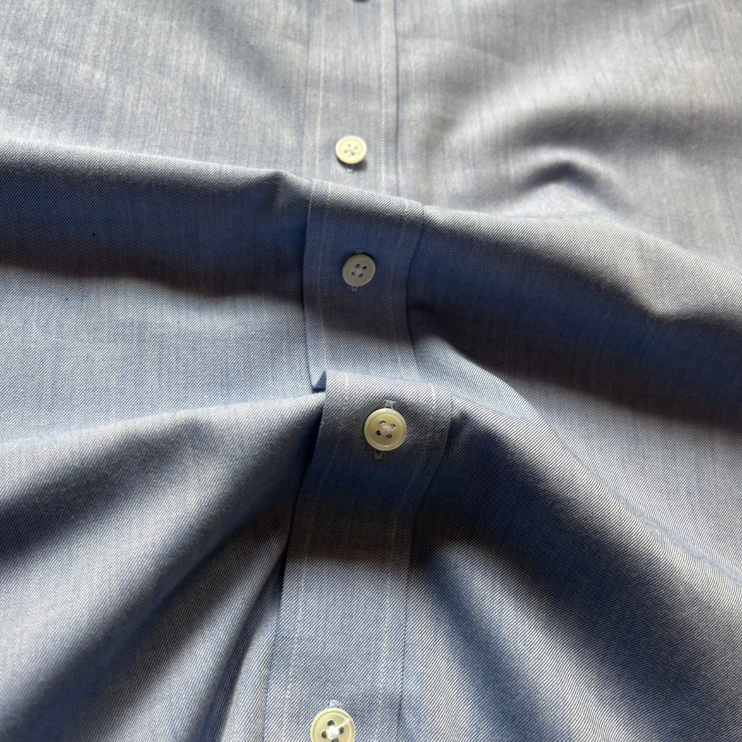 Close-up of a light blue button-down shirt fabric with cream-colored buttons, showing the texture of the cotton weave and the button placket.