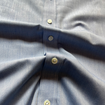 Close-up of a light blue button-down shirt fabric with cream-colored buttons, showing the texture of the cotton weave and the button placket.