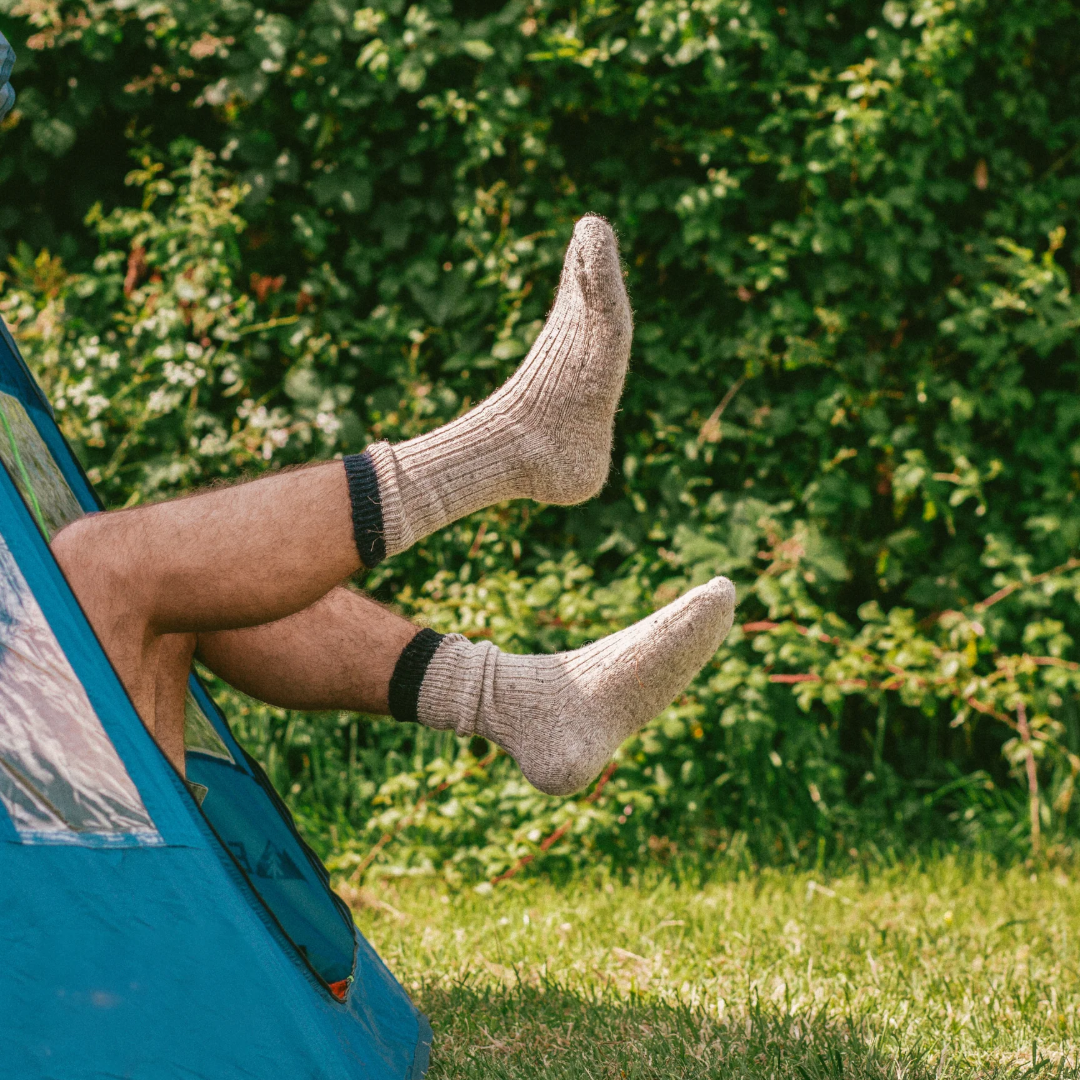 A person's legs and feet wearing beige ribbed knit socks, sitting inside a blue tent in a grassy outdoor setting with a green hedge in the background.