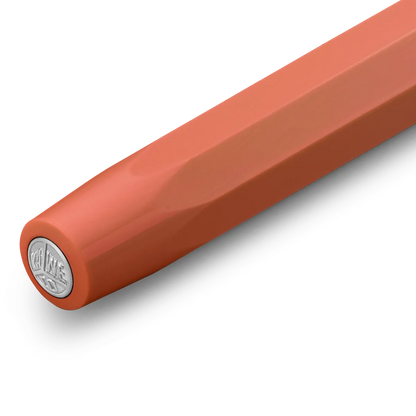 A close-up of a smooth, matte orange-brown cylindrical product, likely a pen or stylus, showing a silver circular logo stamp on the end cap against a clean white background.