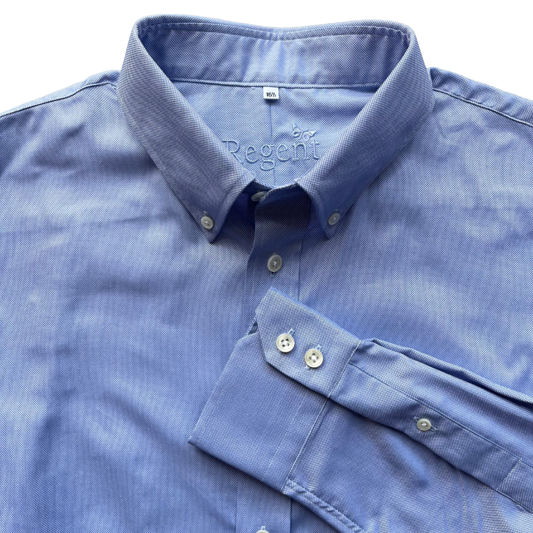 A close-up of a blue long-sleeve dress shirt with a spread collar and folded cuff, showing the button placket and inner neck label.