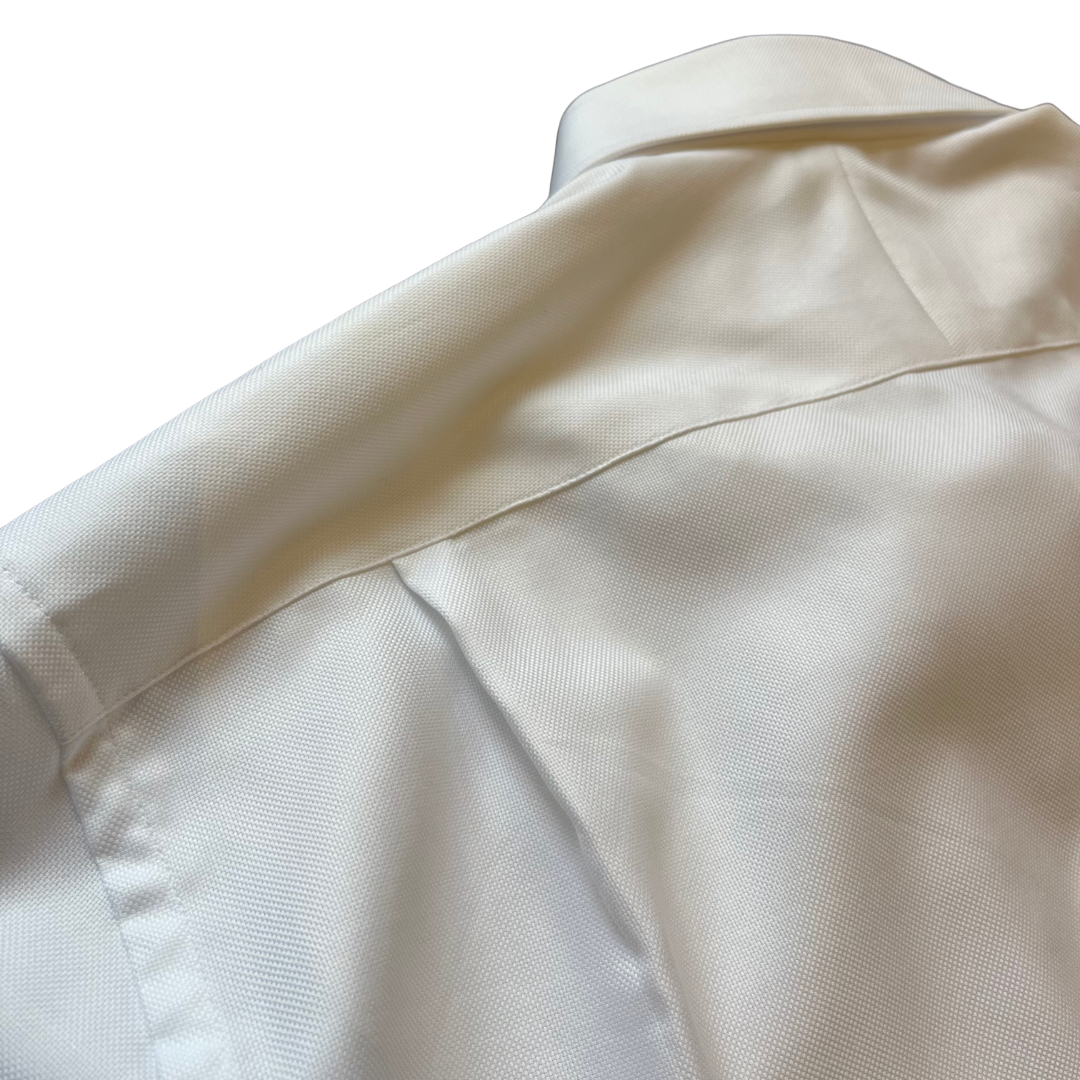 Close-up of a white or off-white shirt fabric showing the collar, shoulder seam, and fine woven texture against a plain white background.