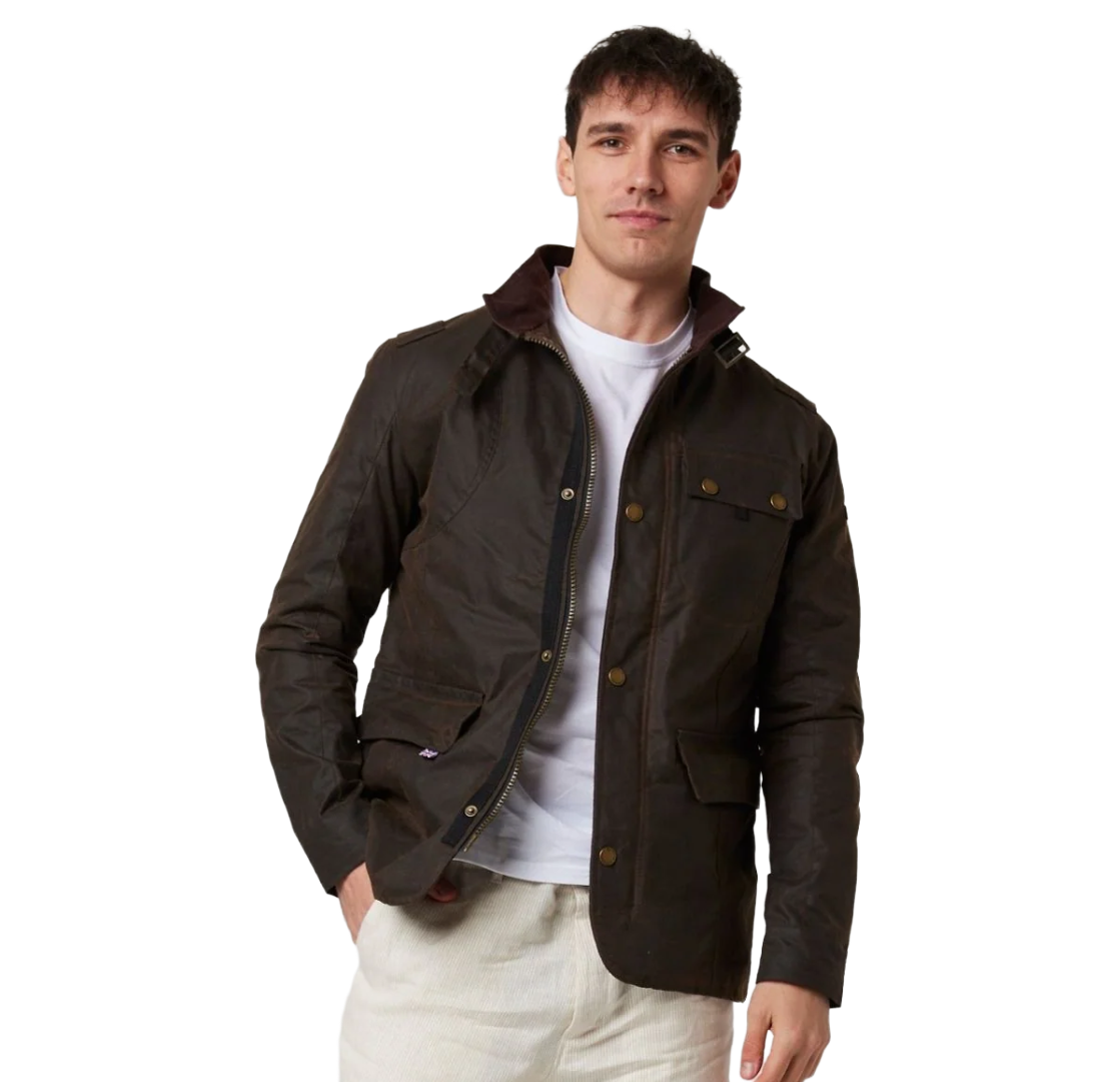 A man wearing a dark brown waxed cotton utility jacket with a white t-shirt and off-white trousers, standing against a solid white background.