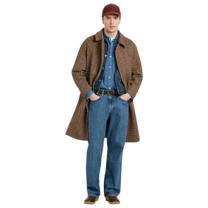 A man modeling a brown and tan houndstooth overcoat, layered over a blue denim shirt and jeans, wearing a maroon baseball cap and brown shoes, set against a plain white background.
