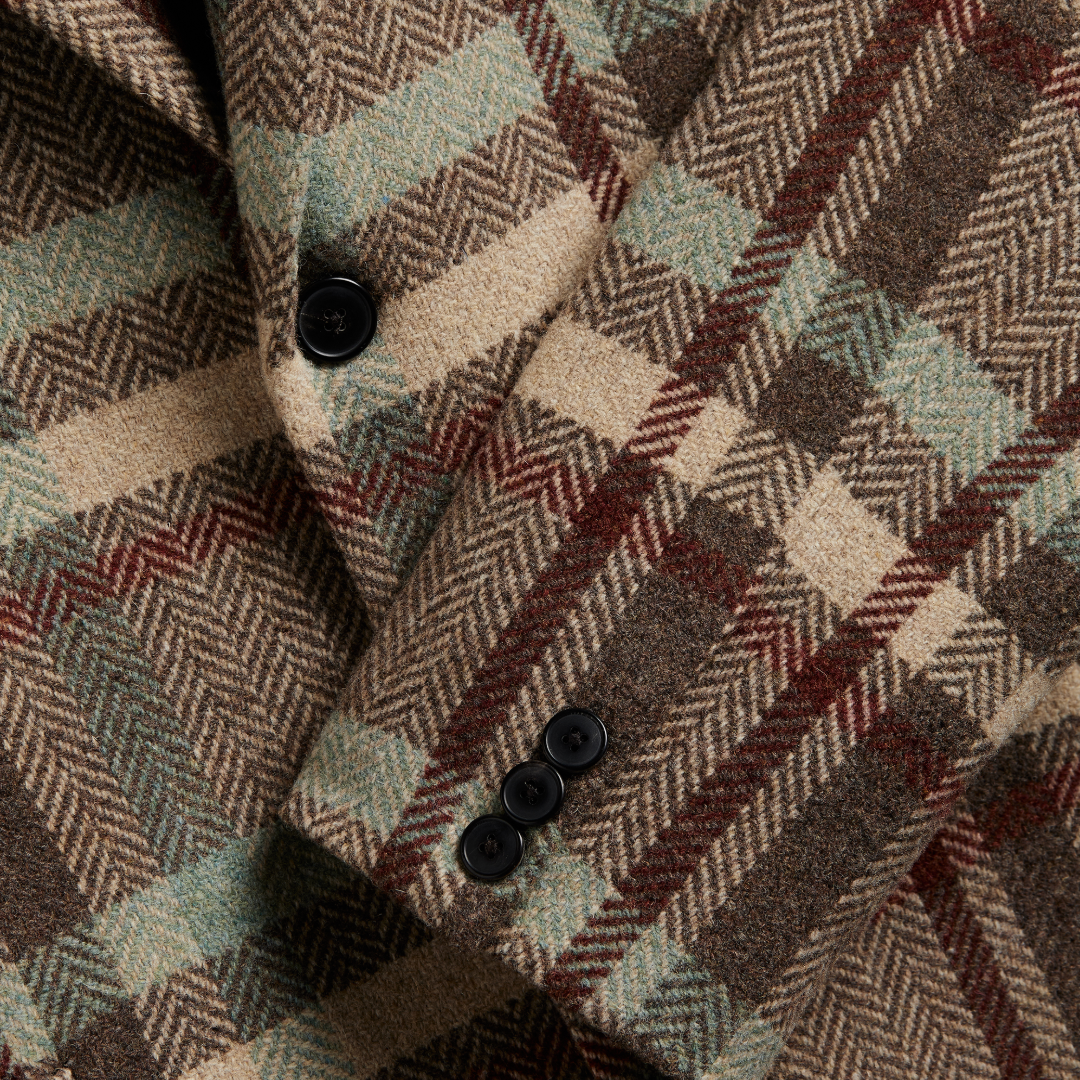 Close-up of a plaid wool coat fabric with a herringbone weave, featuring shades of brown, beige, muted green, and burgundy, accented by black buttons.