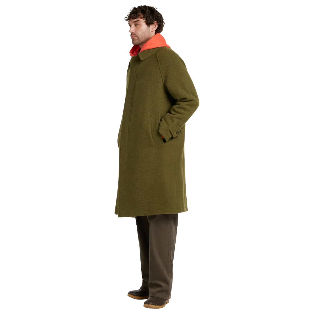 A man wearing a mid-length olive green wool coat with a bright orange collar, paired with brown trousers and dark shoes, shown in profile against a plain white background.
