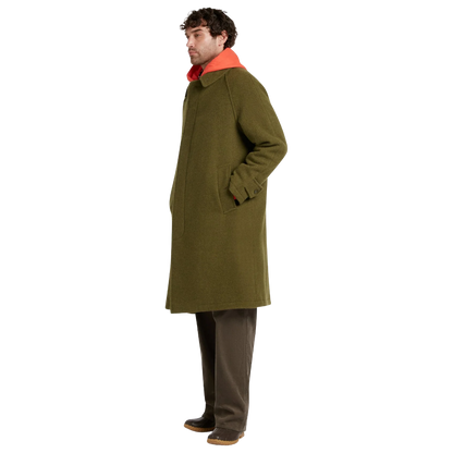 A man wearing a mid-length olive green wool coat with a bright orange collar, paired with brown trousers and dark shoes, shown in profile against a plain white background.