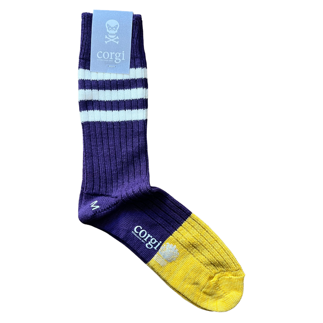 A single crew sock with a purple and yellow color-blocked design, featuring light blue and white stripes on the leg, a yellow toe cap, and a light blue paper tag at the top with a corgi skull logo and the brand name 'corgi'.