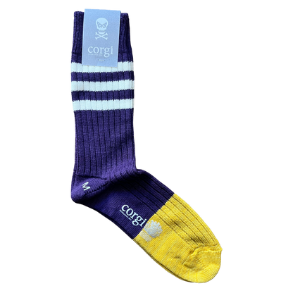 A single crew sock with a purple and yellow color-blocked design, featuring light blue and white stripes on the leg, a yellow toe cap, and a light blue paper tag at the top with a corgi skull logo and the brand name 'corgi'.