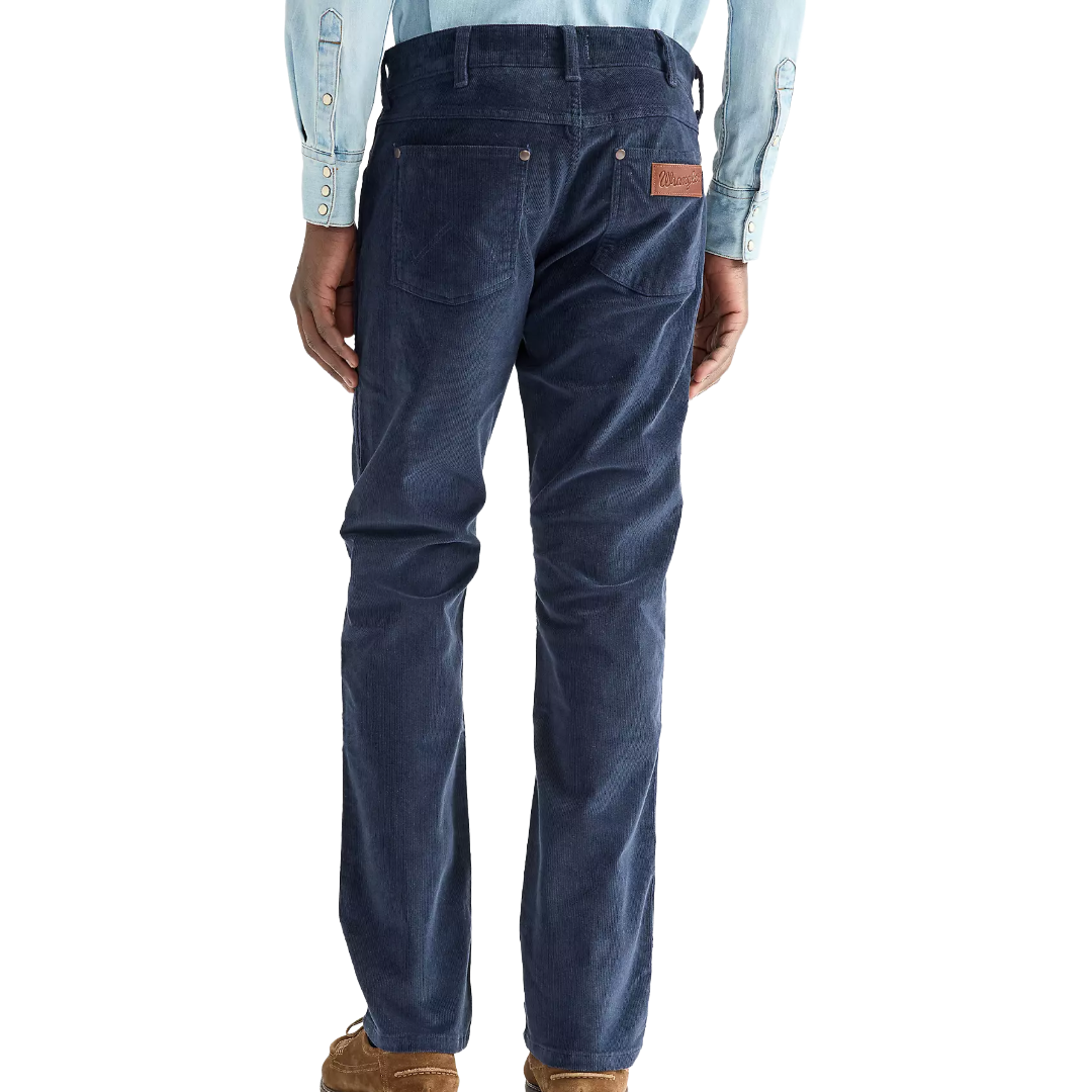 Rear view of a person wearing dark blue corduroy pants with a brown leather patch on the back pocket, paired with a light blue shirt and brown shoes, against a white background.
