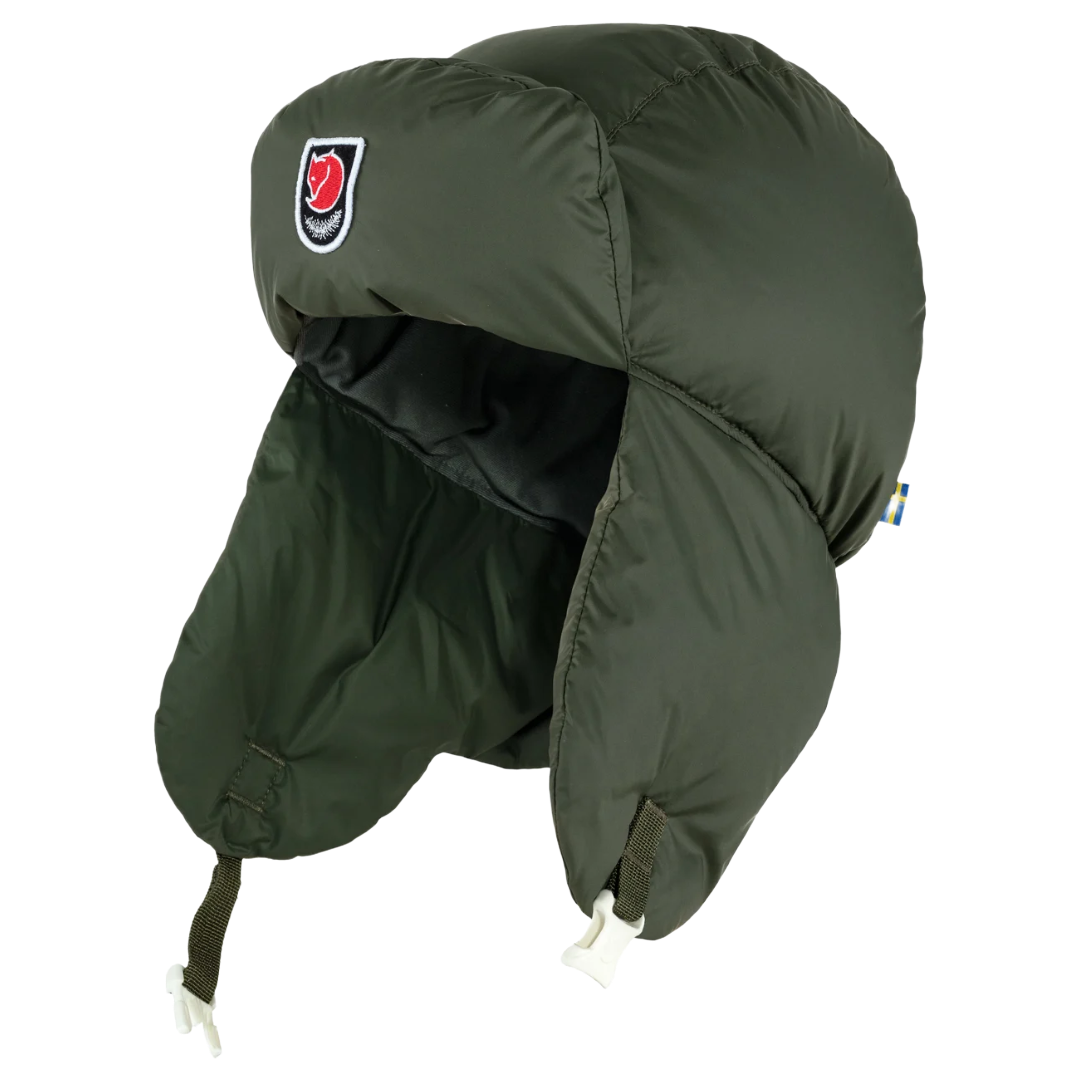 An olive green padded military-style trapper hat with ear flaps and chin straps, featuring a red and black shield logo on the side, shown against a plain white background.