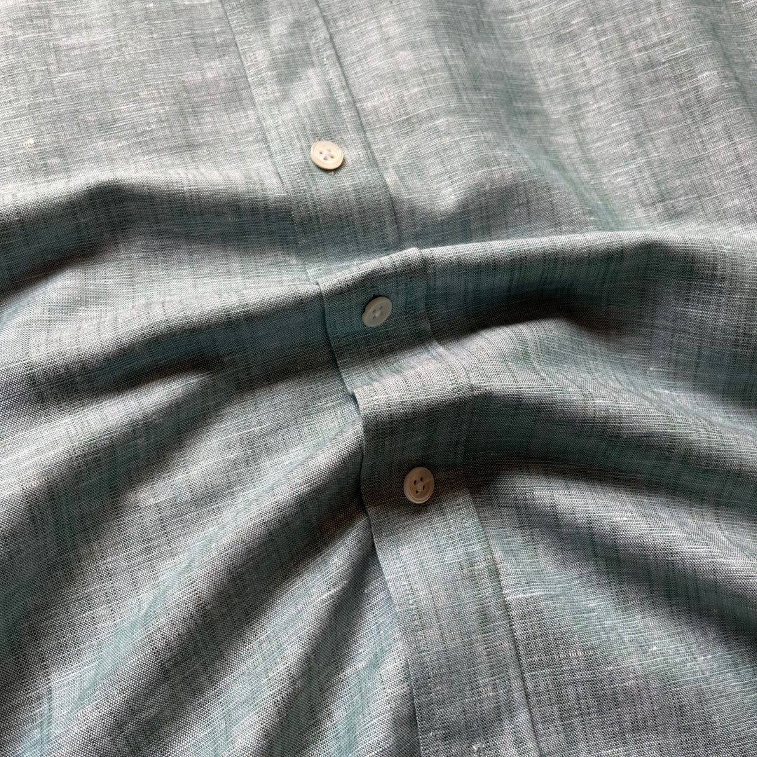 Close-up of a textured linen shirt in a muted sage green color, showing the fabric weave and three buttons.