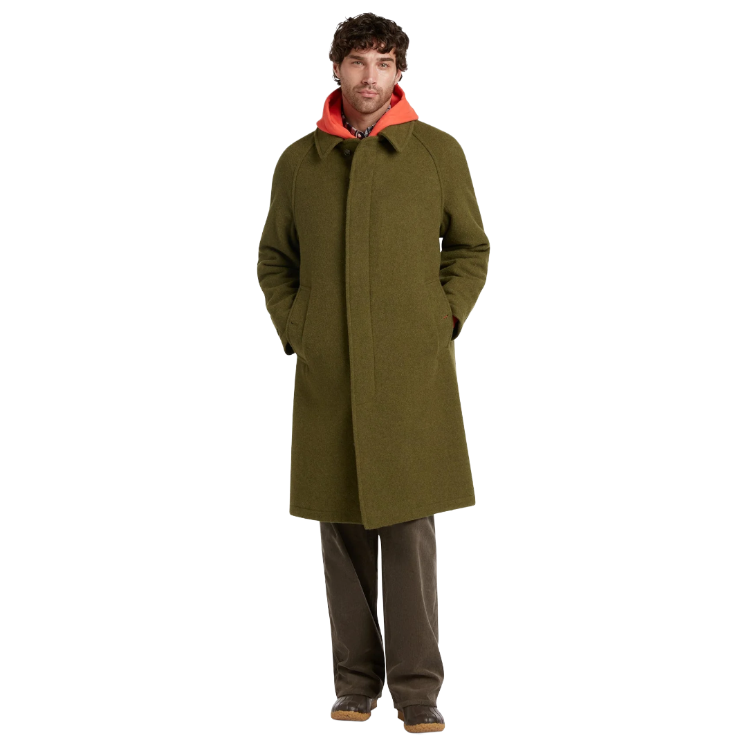 A man wearing a long olive green wool-blend coat over a bright orange hooded sweatshirt, paired with dark brown trousers and brown shoes, standing against a solid white background.