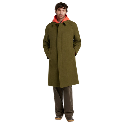 A man wearing a long olive green wool-blend coat over a bright orange hooded sweatshirt, paired with dark brown trousers and brown shoes, standing against a solid white background.