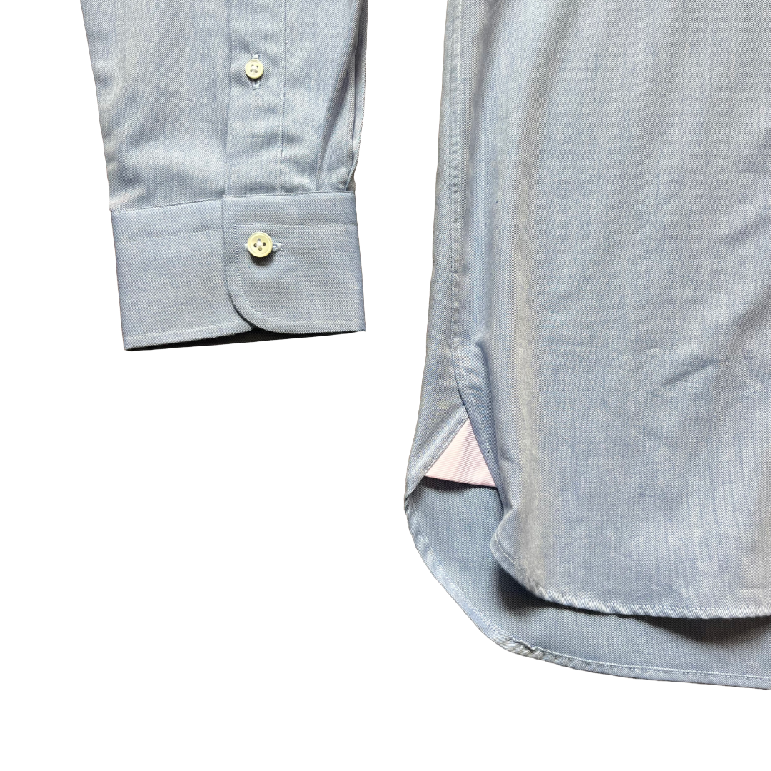 Close-up detail shot of a light blue chambray button-down shirt, showing the folded cuff with a button and the side slit hem.