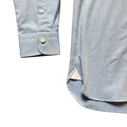 Close-up detail shot of a light blue chambray button-down shirt, showing the folded cuff with a button and the side slit hem.