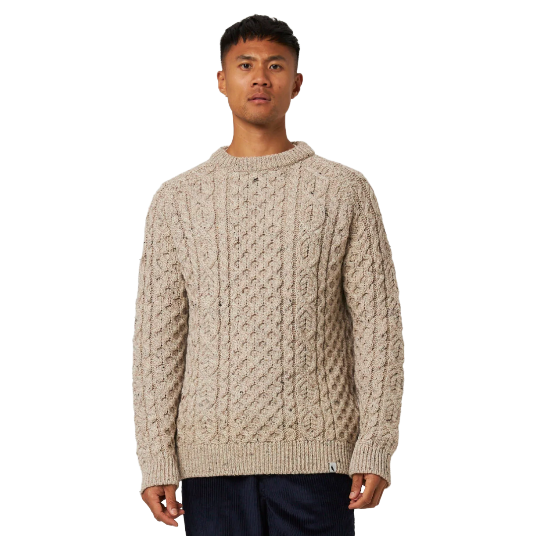 A man wearing a beige cable-knit crewneck sweater, standing against a plain white background.