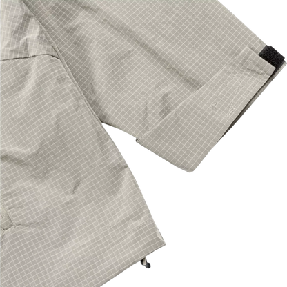 Close-up of a light gray and white grid-patterned garment, showing the fabric texture, a cuff with a black hook-and-loop tab, and a small black adjustment loop near the hem, set against a plain white background.