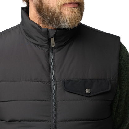 Close-up of a man wearing a black quilted zip-front vest over a dark green knit sweater, with a white studio background.